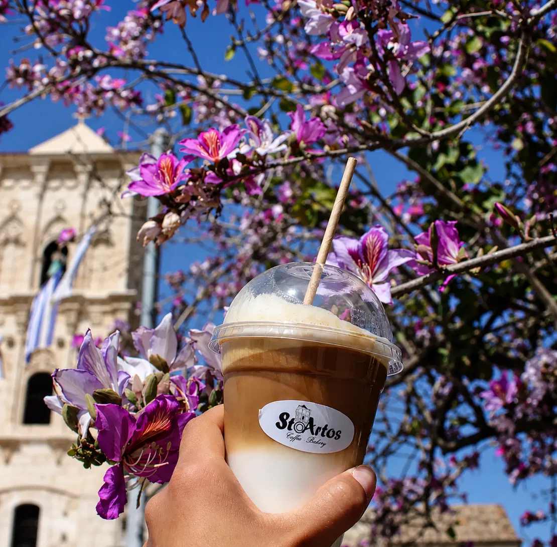 An iced coffee in a StoArtos cup held up in front of the Church of Saint Lazarus, framed by purple bauhinia blossoms